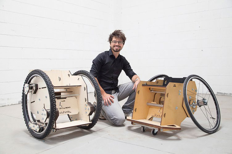 A man kneels beside two prototypes of wooden off-road mobility devices with large wheels, showcasing their design.