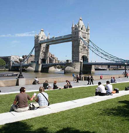 People relax on a grassy area in an urban park, with London's Tower Bridge spanning the river in the background.