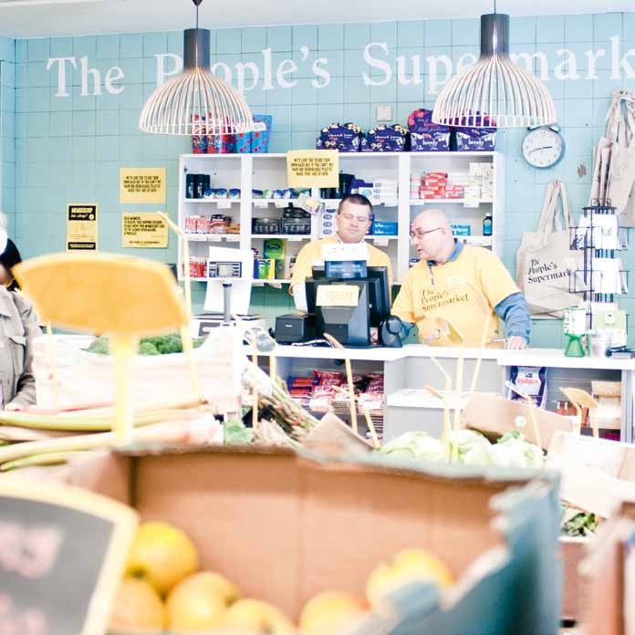Two employees wearing yellow shirts behind the checkout counter of "The People's Supermarket," with fresh produce in the foreground.