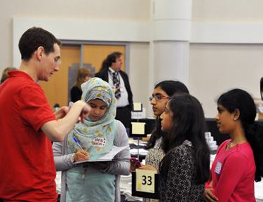 A man in a red shirt talks to four young girls, one of whom is wearing a hijab and writing on a clipboard, at an event.