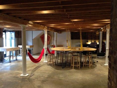 Interior view of an open-plan co-working space featuring large tables, stools, white support columns, and red hammocks.
