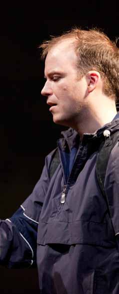 Side profile of a man in a dark blue jacket and backpack, looking down thoughtfully, possibly an actor in costume.