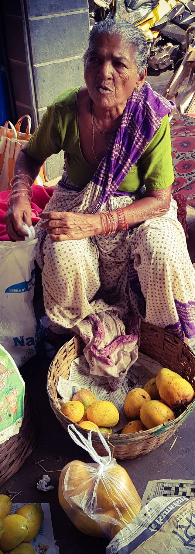 An elderly woman vendor sitting on the ground with baskets of fresh mangoes and other produce at a market.