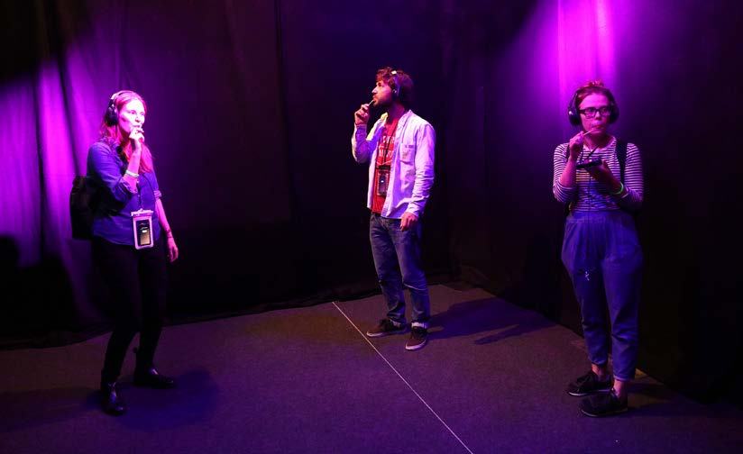 Three individuals, wearing headphones and holding devices, stand in a purple-lit room, participating in an interactive experience.