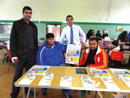 Four men smiling behind a table with leaflets at an indoor community event, with one man holding up a flyer.