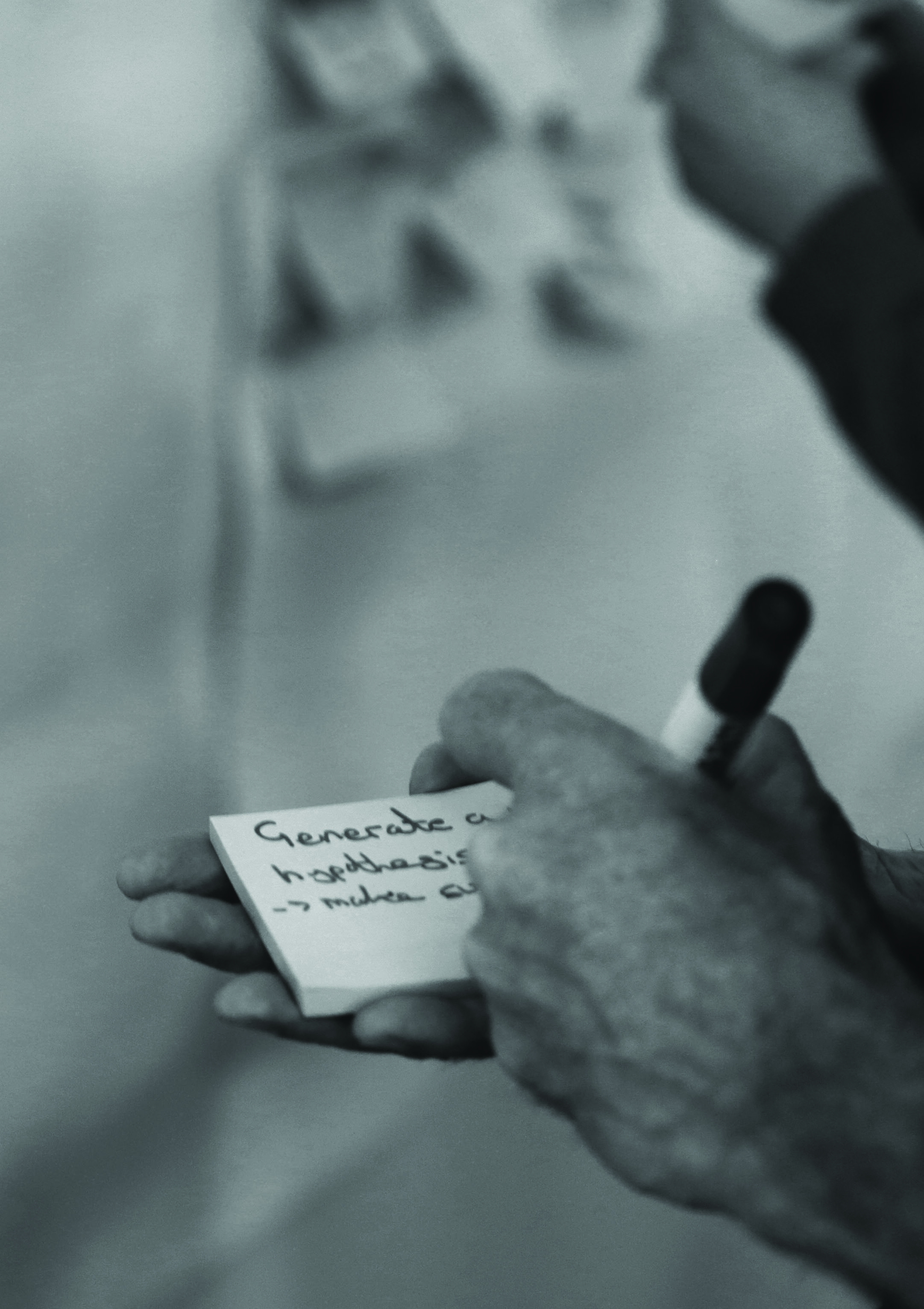 Hands writing "Generate a hypothesis" on a sticky note with a black marker.