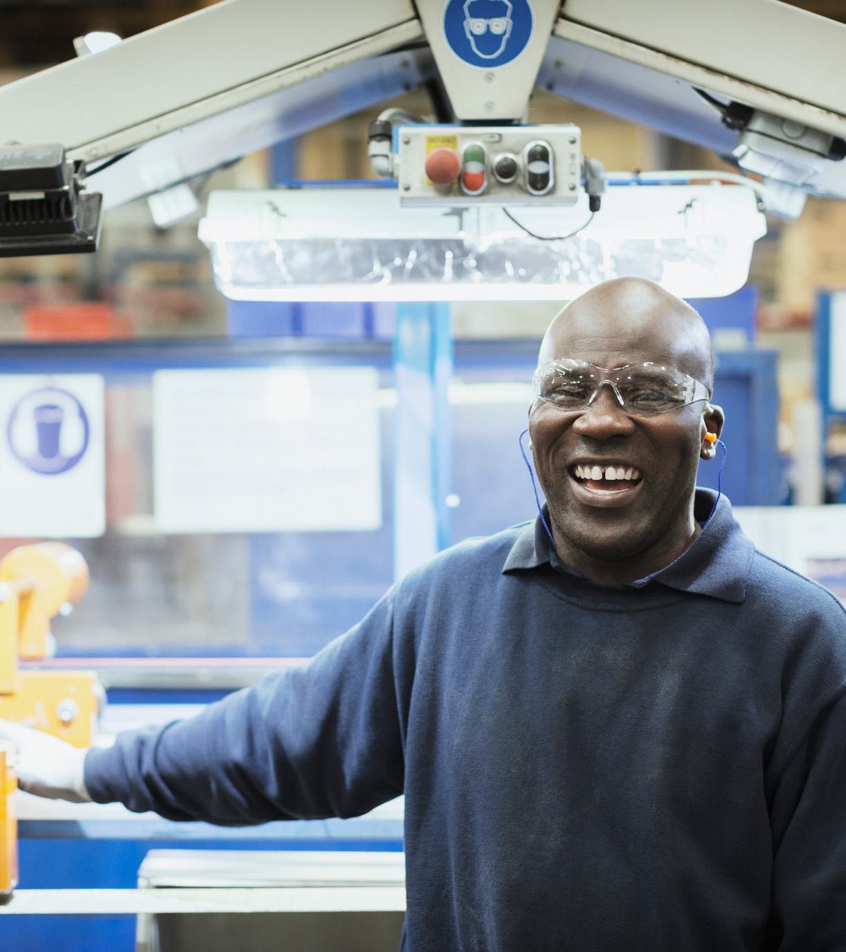 Smiling male worker wearing safety glasses and earplugs in a factory setting with industrial machinery behind him.