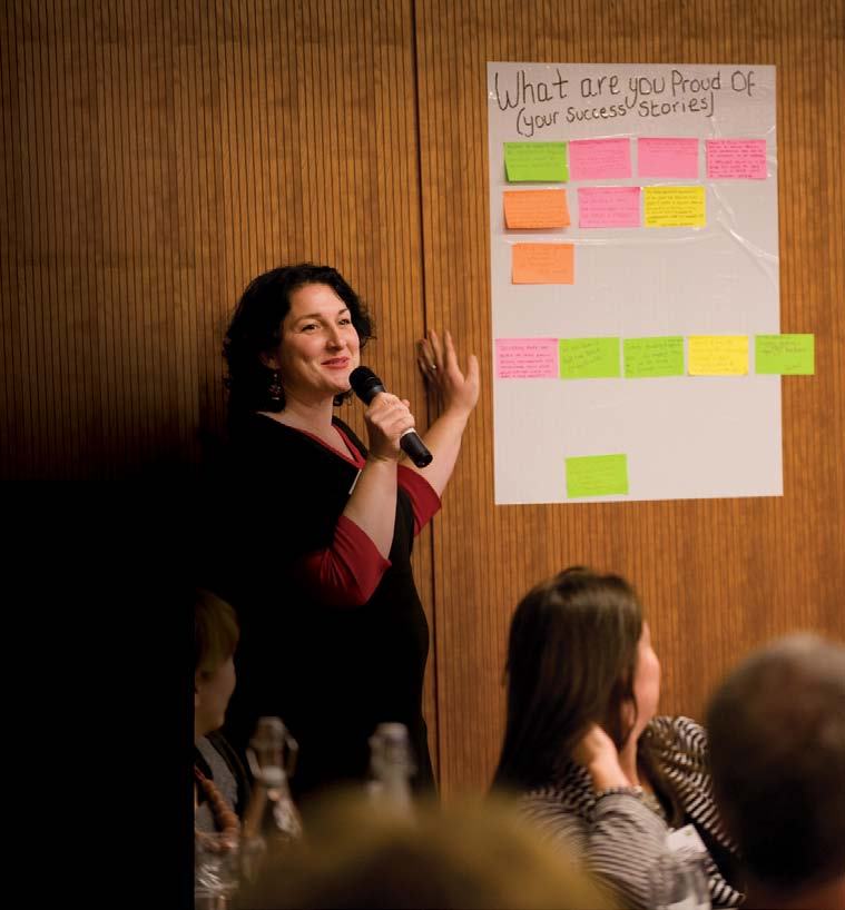 A woman speaks into a microphone next to a whiteboard with sticky notes under the heading "What are you Proud Of (Your Success Stories)".