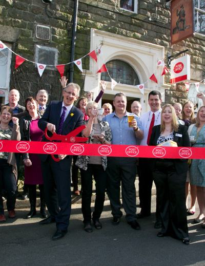 Ribbon-cutting ceremony for a Post Office, with a man cutting a red ribbon using oversized scissors, surrounded by smiling people and bunting.