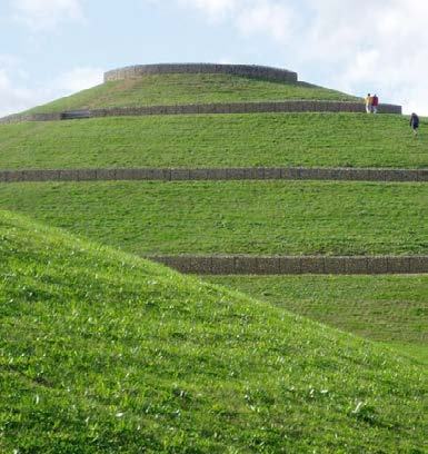 A large, tiered grassy mound with gabion walls and a circular stone structure at the top, under a blue sky.