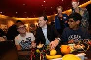 An adult man interacts with two students at a table with fruit, wires, and electronic components, likely during a workshop.