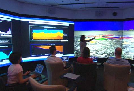 A person interacting with a large 3D city model on a multi-screen data visualization wall in a control room, with several data charts also displayed.