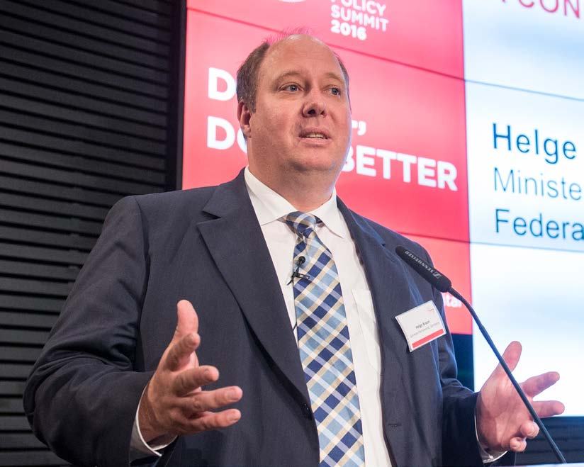 A man in a suit speaks at a podium, gesturing with hands, with a screen behind him showing "POLICY SUMMIT 2016" and "Helge Minister".