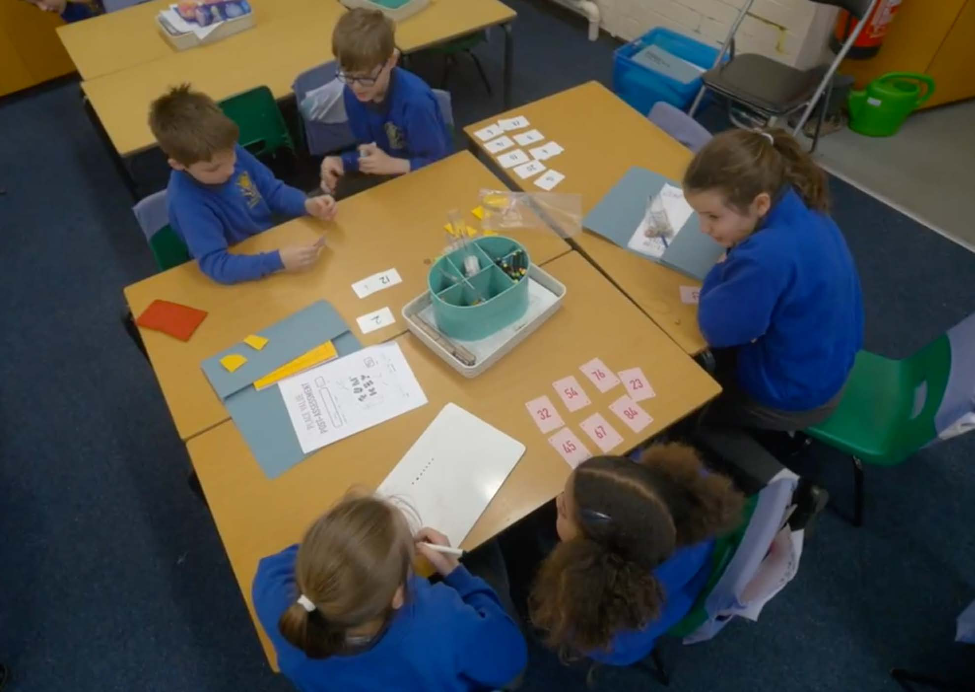 Four children in a classroom working on a math activity at their desks with numbered cards and papers.
