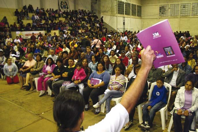 A large audience attends an event, with a speaker or organizer in the foreground holding up a purple brochure.