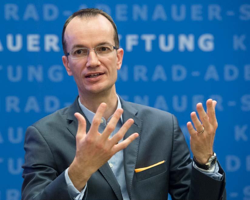 A man in a suit and glasses gestures with both hands in front of a blue wall featuring the "Konrad-Adenauer-Stiftung" logo.