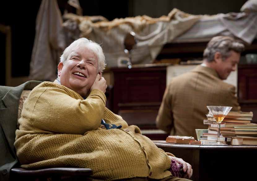 An older man with white hair wearing a yellow cardigan laughs heartily, seated in a chair, while another man plays piano in the background.