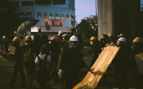 Group of people wearing hard hats and masks, some holding shields, on a street at dusk.