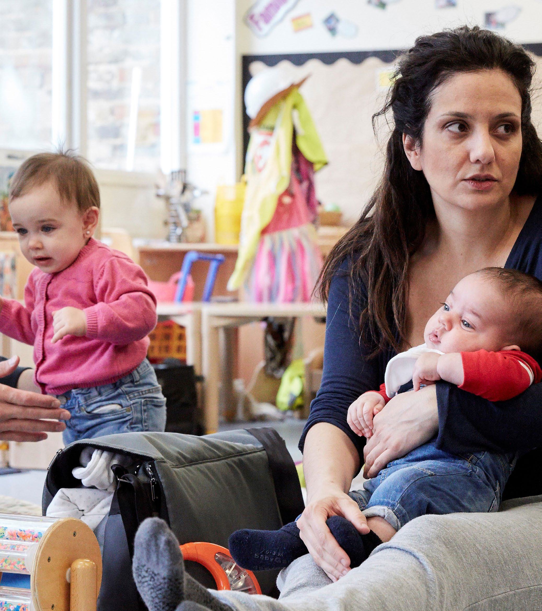 A woman holding an infant, with a toddler in the background, in a room with play equipment.