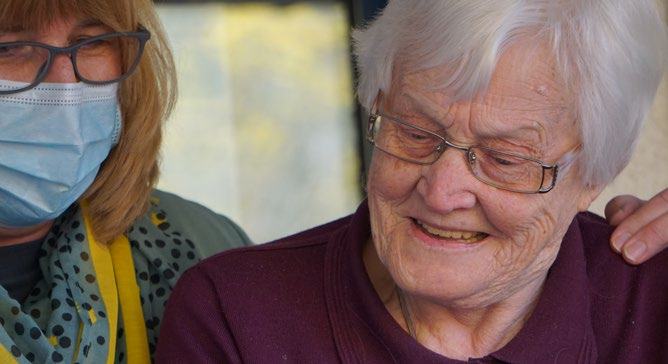 Older woman smiling while another person, wearing a mask, gently touches her shoulder, suggesting care or companionship.