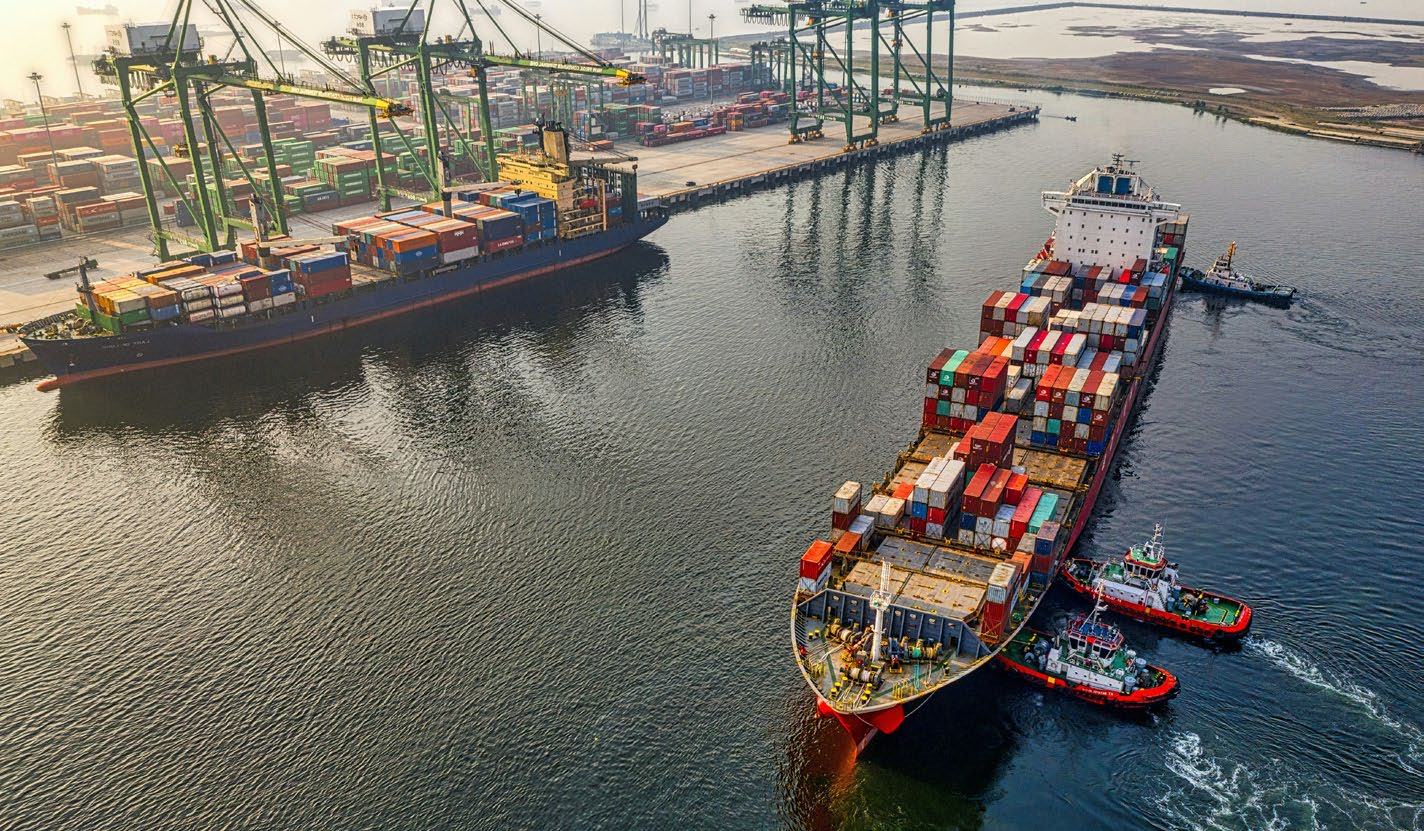 Aerial view of a busy shipping port with two large container ships, one being assisted by tugboats, surrounded by shipping containers and cranes.