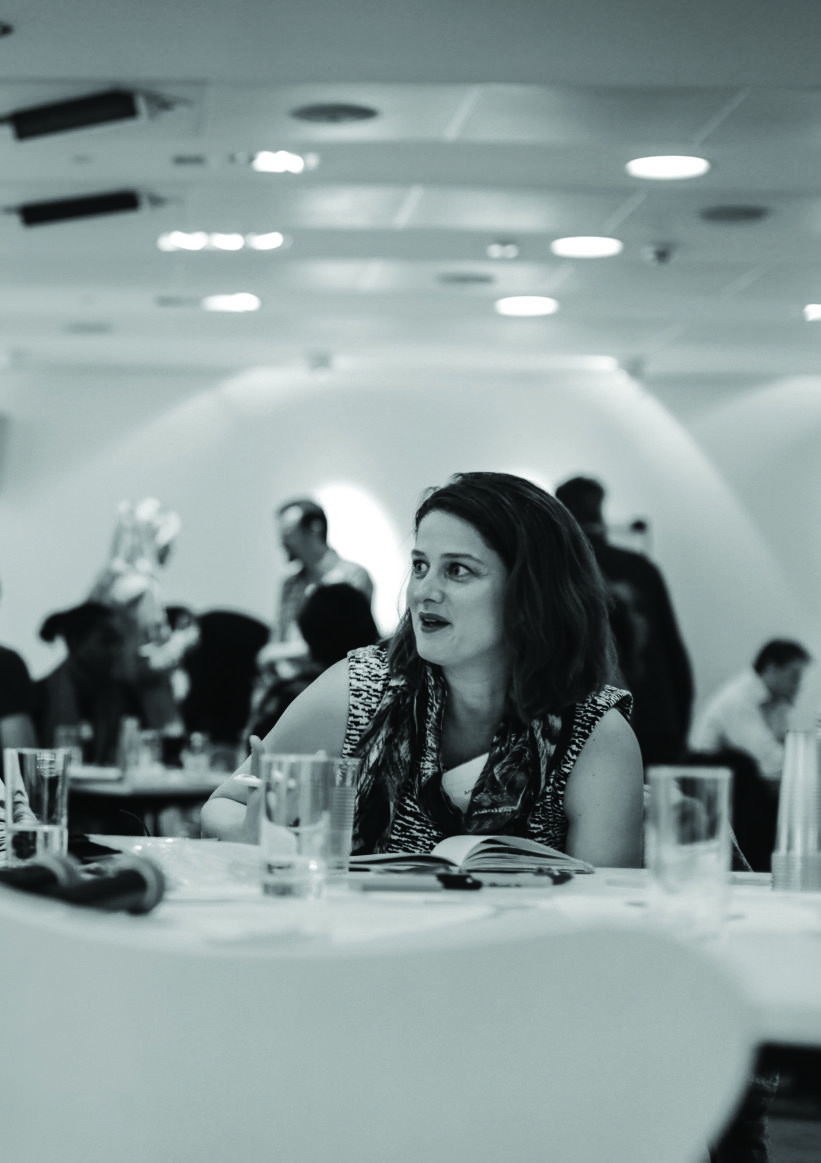 Woman with long hair, wearing a patterned top, seated at a table with an open book, looking engaged in a conference or event setting.