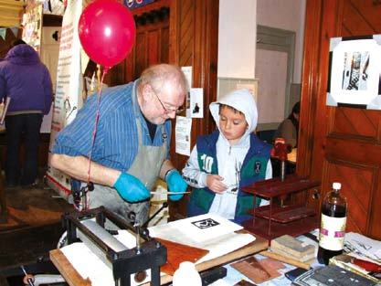 An adult in an apron and gloves demonstrating a printing process to a young boy at an indoor craft event.