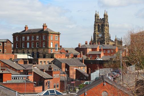 An aerial view of a city with brick buildings and a tall clock tower church in the background, possibly Leeds.