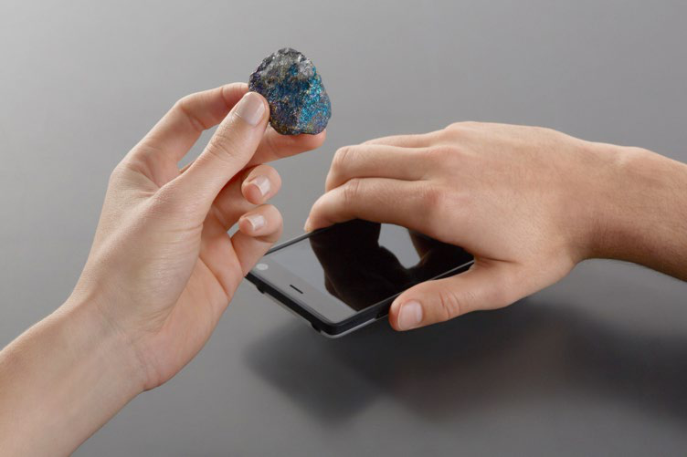 Two hands. One holds an iridescent mineral, the other holds a black smartphone, suggesting interaction or analysis of the object.