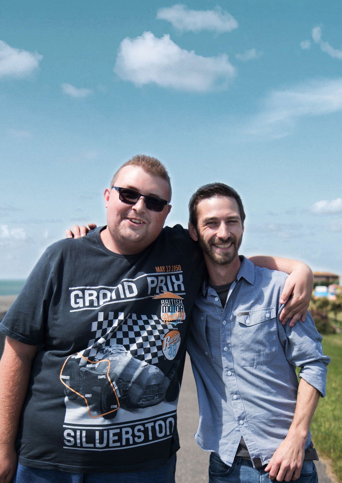 Two men smiling outdoors, one with an arm around the other, under a cloudy blue sky.