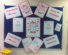 Bulletin board display featuring multiple flyers and signs about a program to "Transform Lives" and "Build recovery," with red smartphone icons.