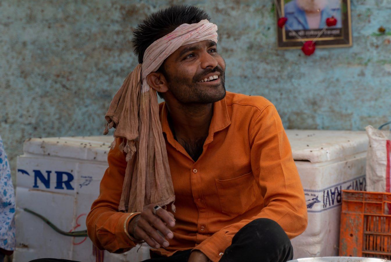 A man with a beard and a head wrap, smiling as he looks upwards, sitting in a market setting with crates behind him.