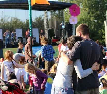 Outdoor community event with a band playing on a stage and a crowd of people, including a couple hugging in the foreground.