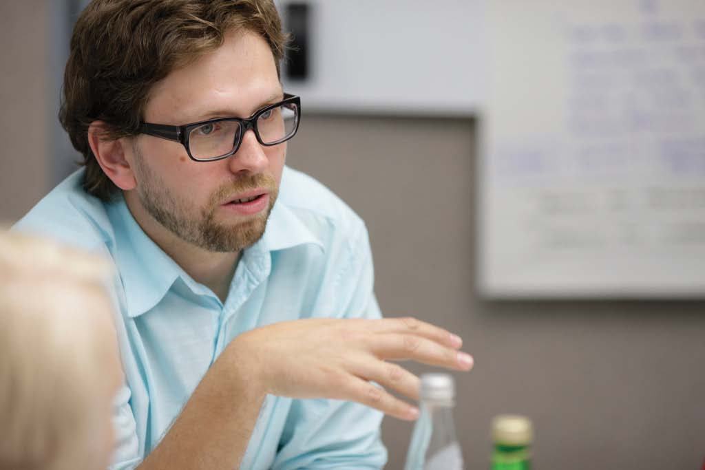 A man with glasses and a beard, wearing a light blue shirt, gestures while speaking in a meeting setting.
