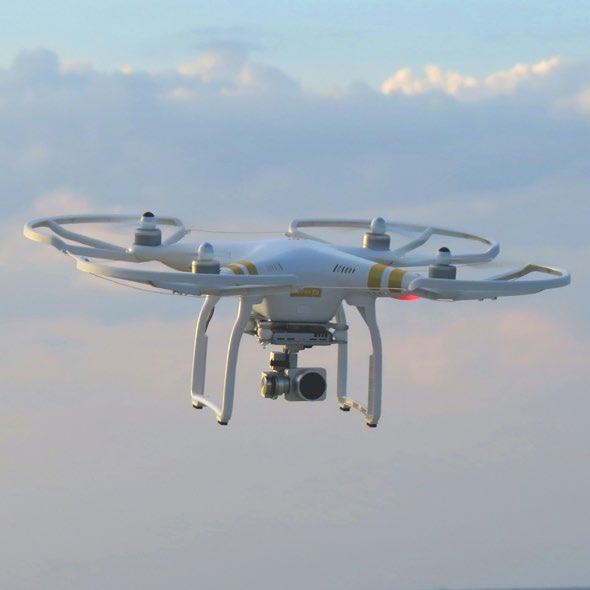 A white drone with four propellers and a camera mounted underneath, flying against a cloudy sky.