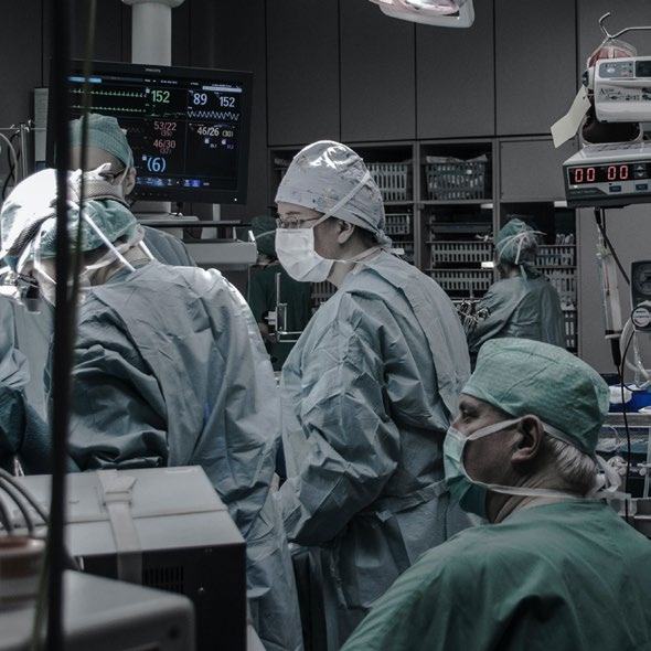 Surgeons in masks and scrubs working in an operating room, with a monitor displaying vital signs in the background.