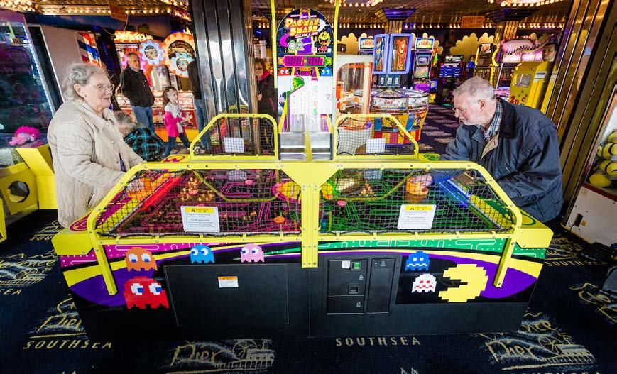 Two elderly people engaging with a colorful arcade game featuring Pac-Man characters in a busy arcade setting.
