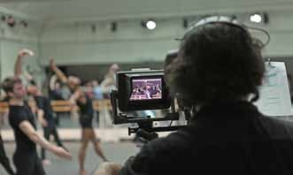 Cameraman filming a group of dancers during a rehearsal, with the camera's display showing the recording.