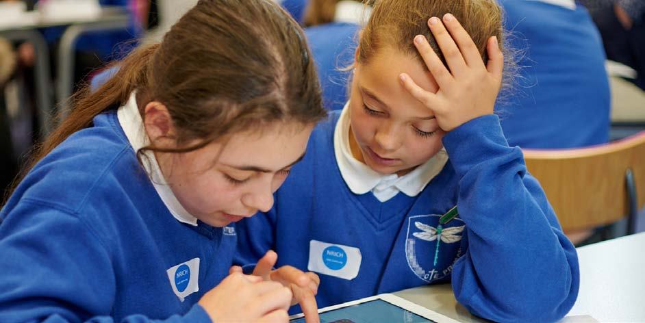 Two schoolgirls in uniform collaborate on a learning activity using a tablet in a classroom setting.