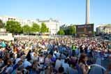 Large crowd gathered outdoors in a city square, surrounded by buildings and a tall tower under a clear blue sky.