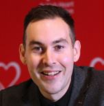 Headshot of a smiling man with short dark hair, wearing a dark jacket and shirt, against a red background.