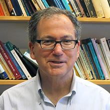 Headshot of a smiling man with glasses and grey hair, with bookshelves visible in the background.