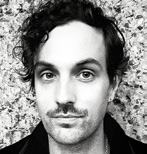 Black and white portrait of a man with dark curly hair and a mustache, looking forward.