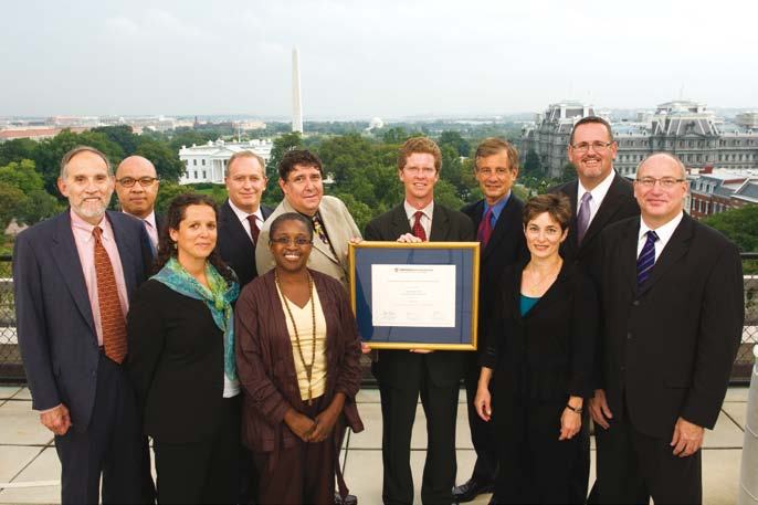 A group of eleven professionals pose on a rooftop with the Washington Monument in the background; one man holds a framed certificate.