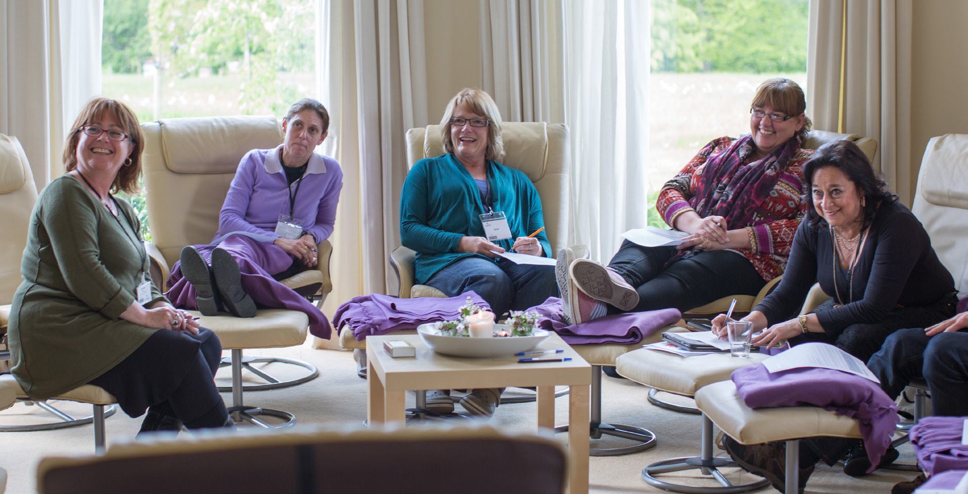 Group of six women seated in a circle during a therapy session or workshop, some taking notes, some smiling.