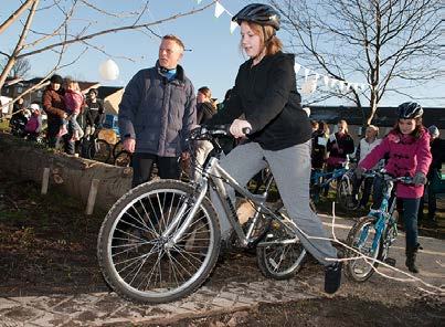 A young woman balances on a bicycle on a dirt path, with an adult observing and other people in the background, likely at a bike event.
