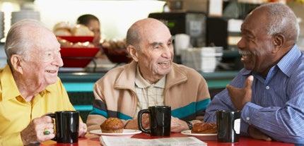 Three elderly men smiling and conversing over coffee and pastries at a cafe.