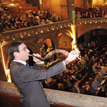 A man in a suit playing a shofar from an upper level in a large hall filled with an audience, likely during a religious or cultural ceremony.