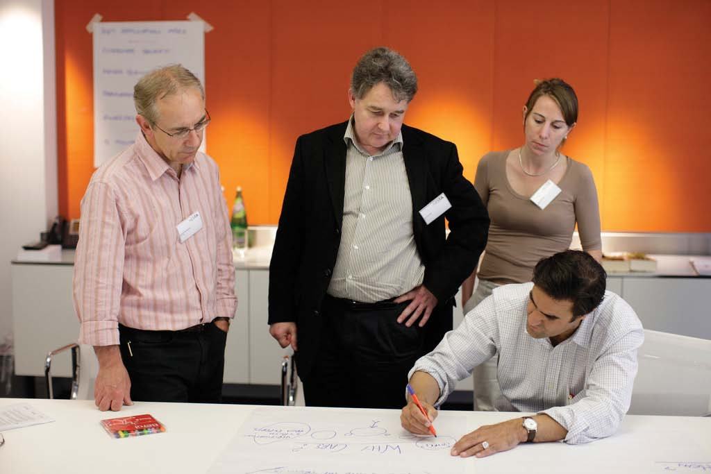 A group of four professionals in a meeting room, observing a colleague writing on a large paper during a collaborative session.