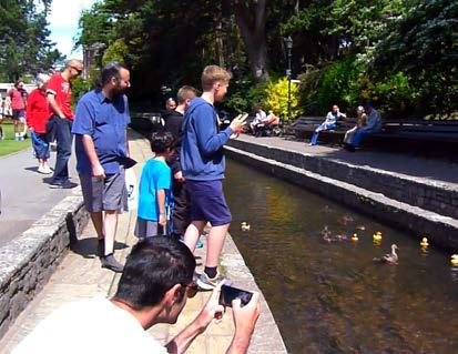 People gathered along a canal watching a rubber duck race, with some taking photos.